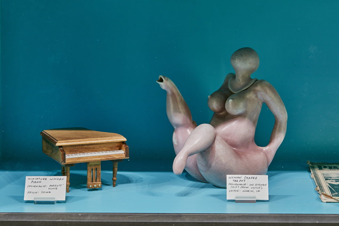 a closeup of the middle display, showing a miniature wooden piano and a teapot shaped like a nude woman