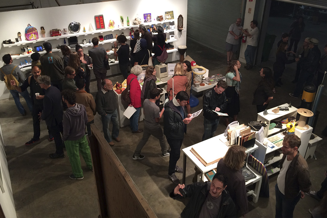 A crowd in a gallery, viewed from above, surrounded by shelves containing seemingly random objects. Some are using their phones to scan tags beneath each object, and others are holding information sheets
