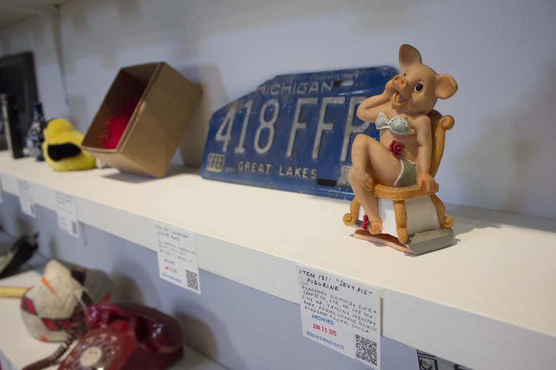 Closeup of objects on shelves, showing a ceramic figurine of a bikini-wearing pig, next to a paper tag with its name, provenance, date it was archived, and a QR code
