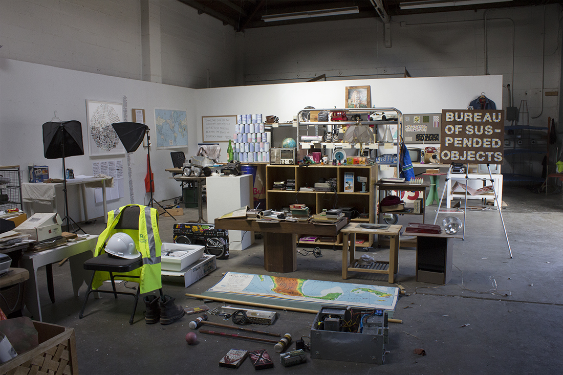Messy studio space where found objects fill found shelves and tables, and are also laid out on the floor. In the background is a photographic setup with box lights and a desk with an office chair.