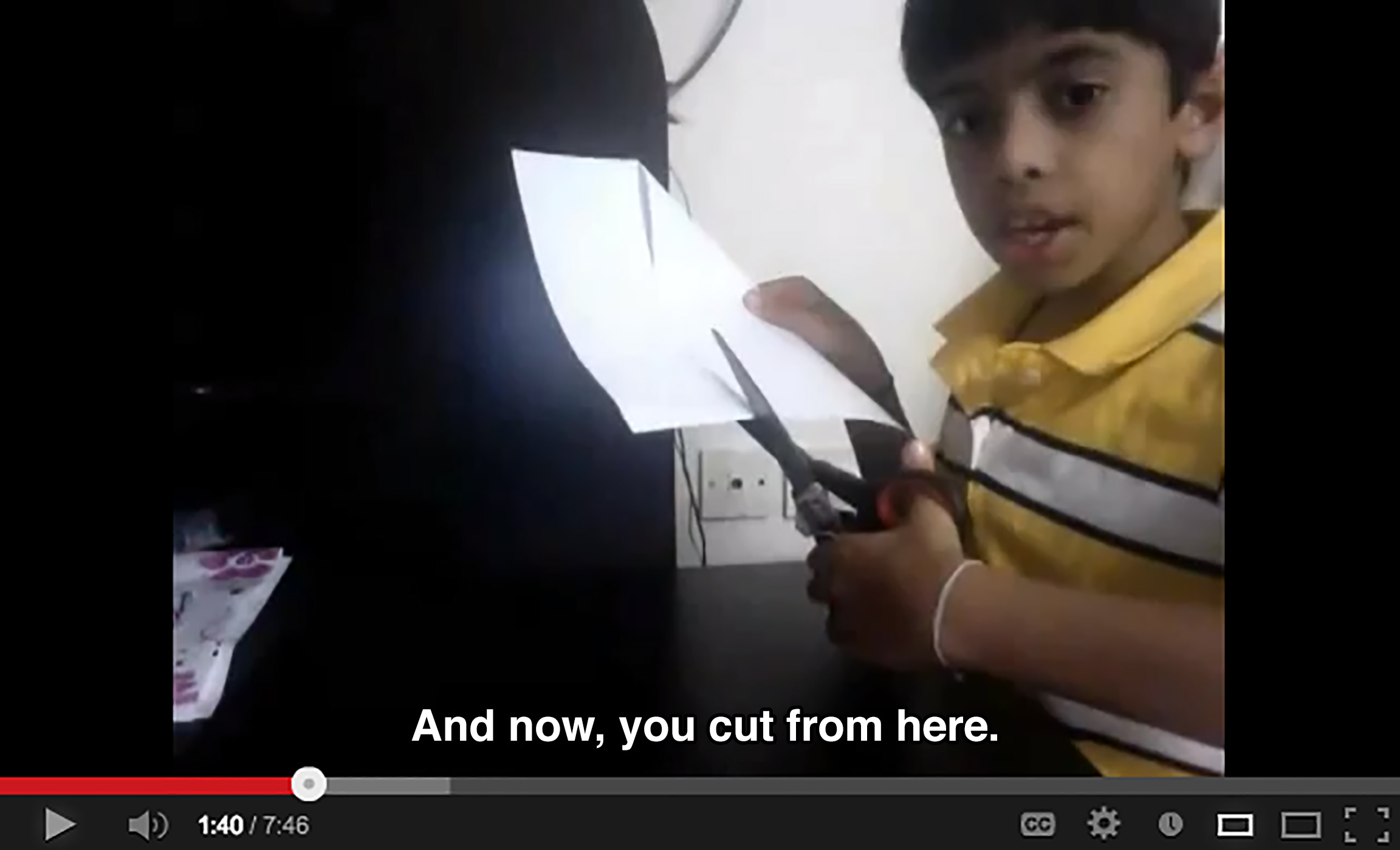 A young boy sits at a desk and cuts paper with scissors while looking at the camera, saying: And now, you cut from here.