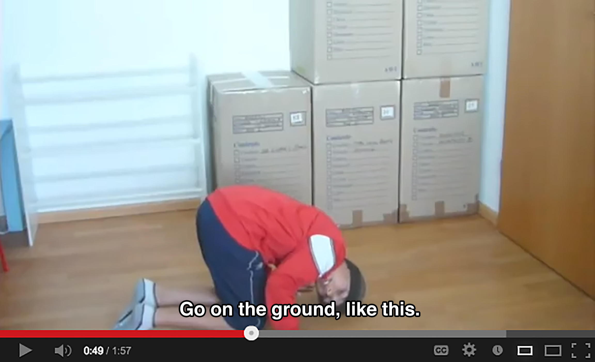 A boy has his knees, hands, and head on the floor in a room with an empty shoe rack and stacked cardboard boxes. He says: Go on the ground, like this.