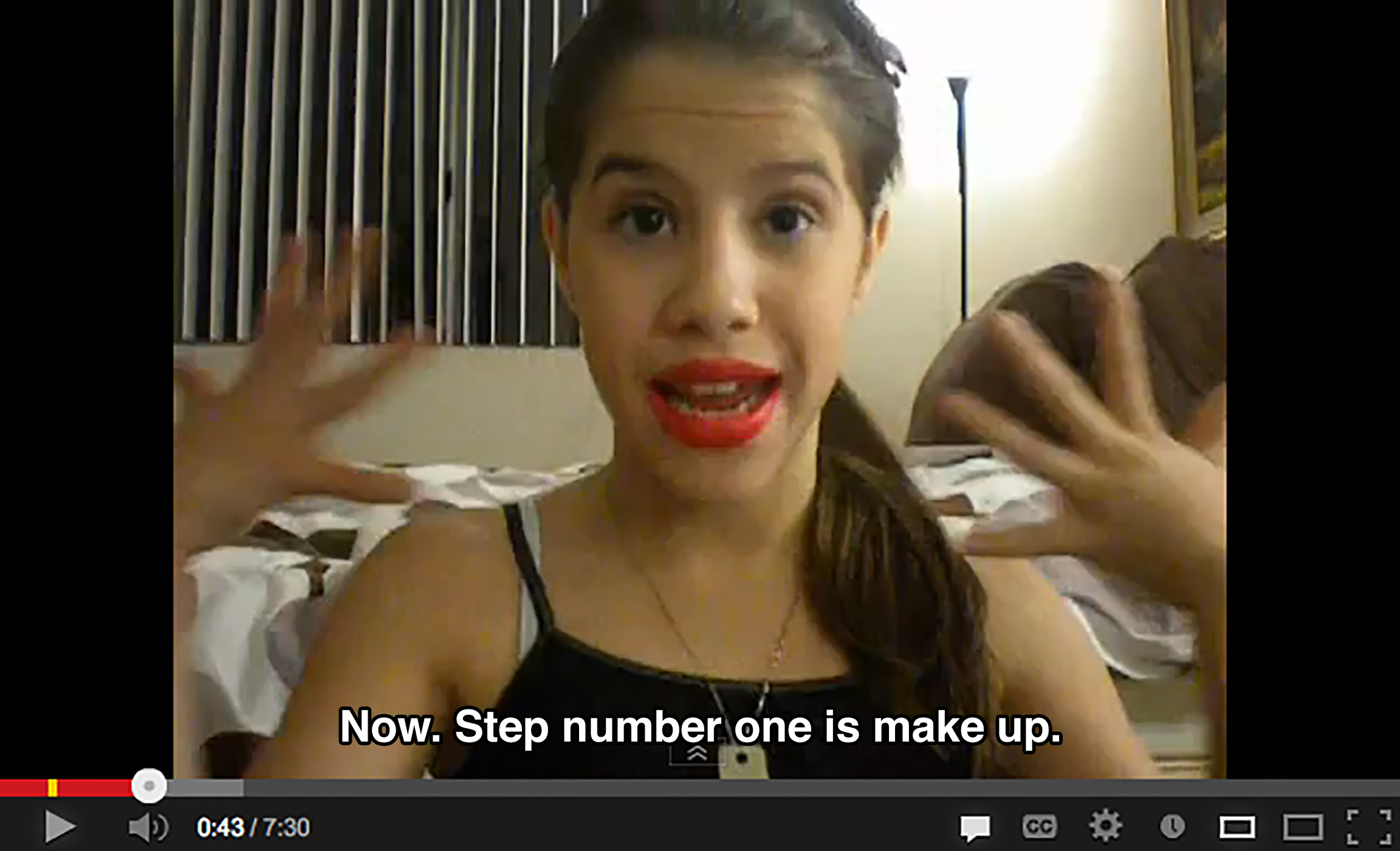 A young girl with a lot of red lipstick on in a bedroom gestures at her face and says: Now. Step number one is make up.