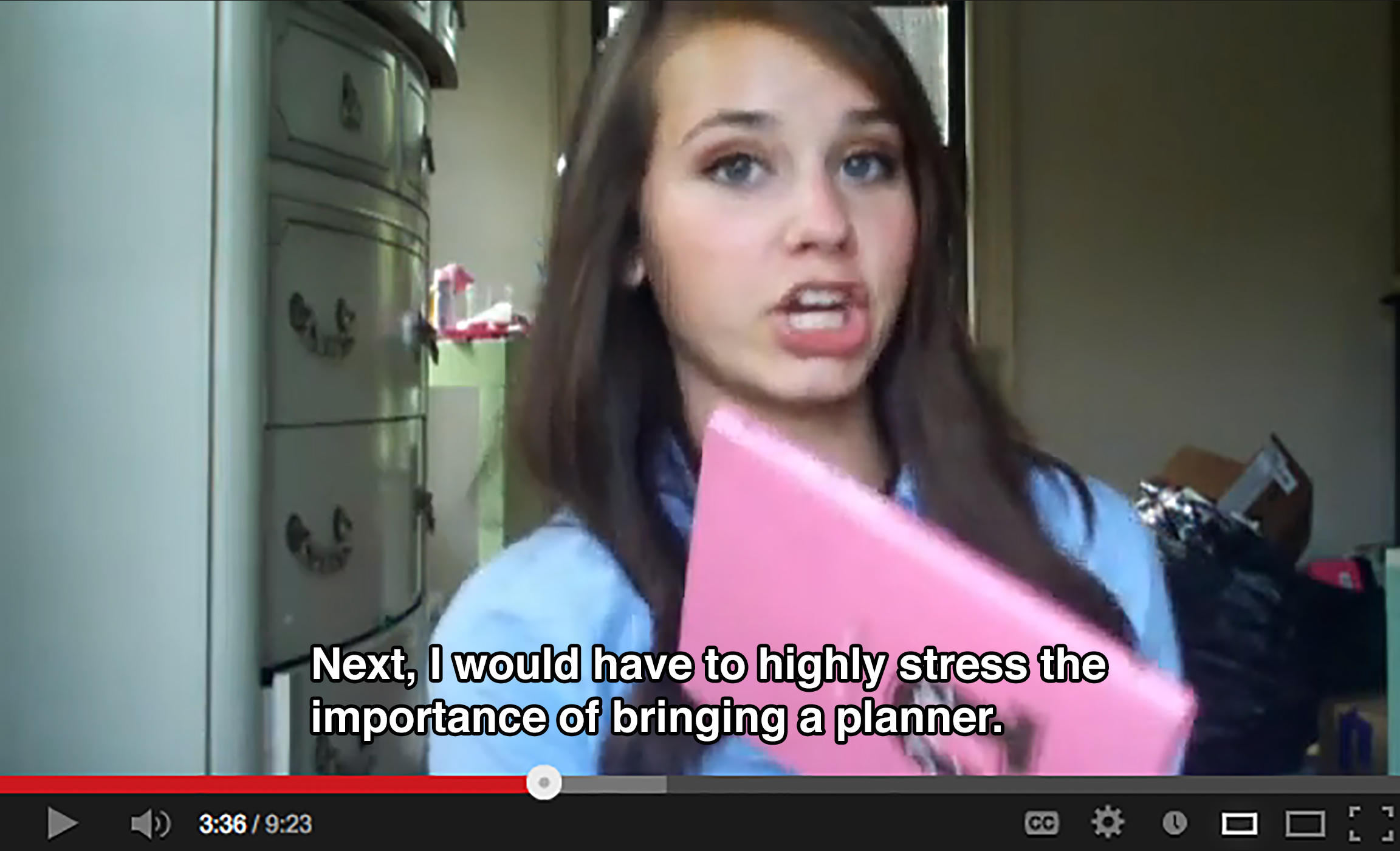 A teenager in a messy bedroom holds up a pink planner to the camera and says: Next, I would have to highly stress the importance of bringing a planner.