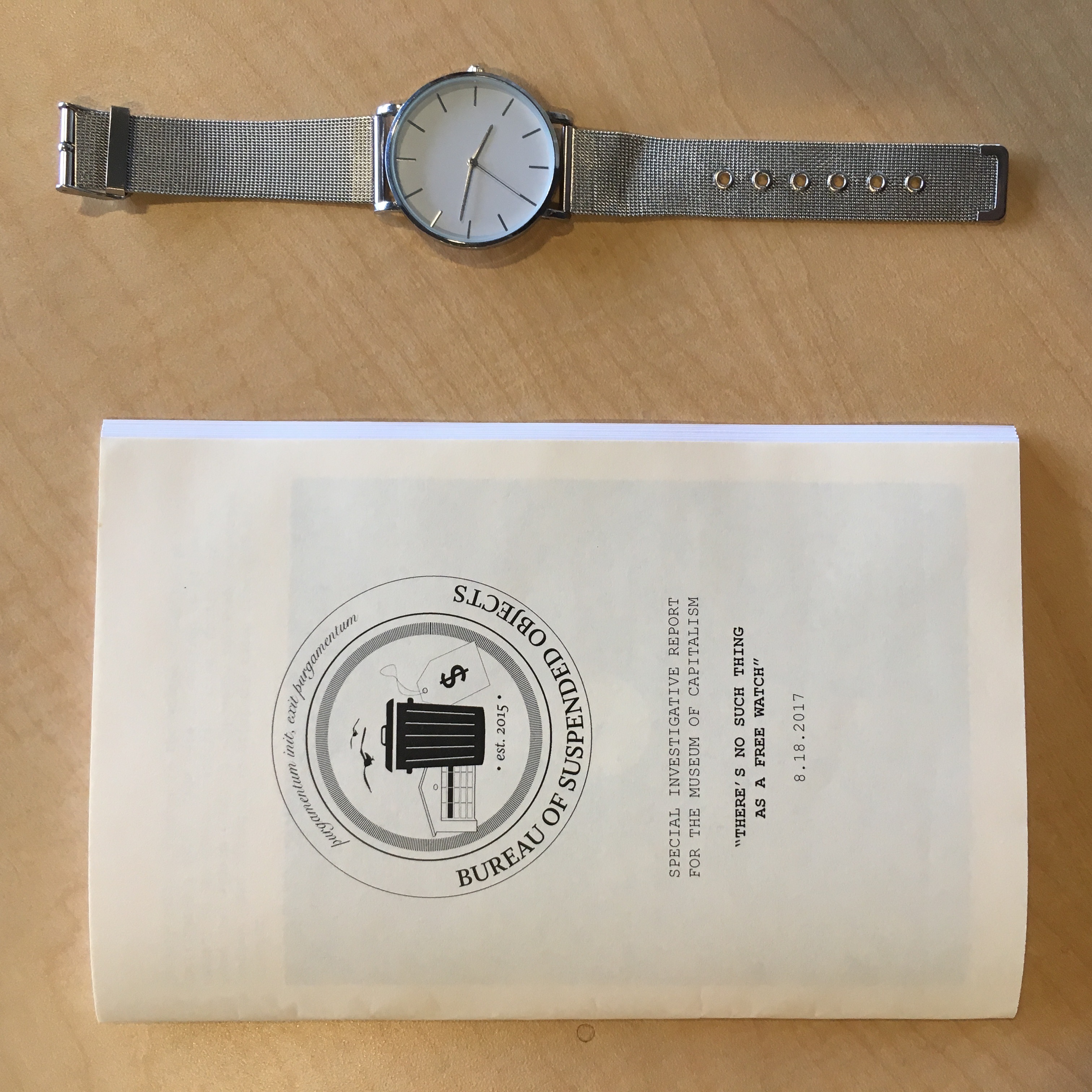 The silver watch next to a zine with the logo of the Bureau of Suspended Objects and the title: There's No Such Thing as a Free Watch