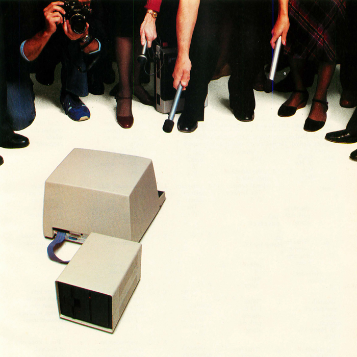 The feet of a crowd of reporters with a hand holding a microphone down toward a computer as if to interview it