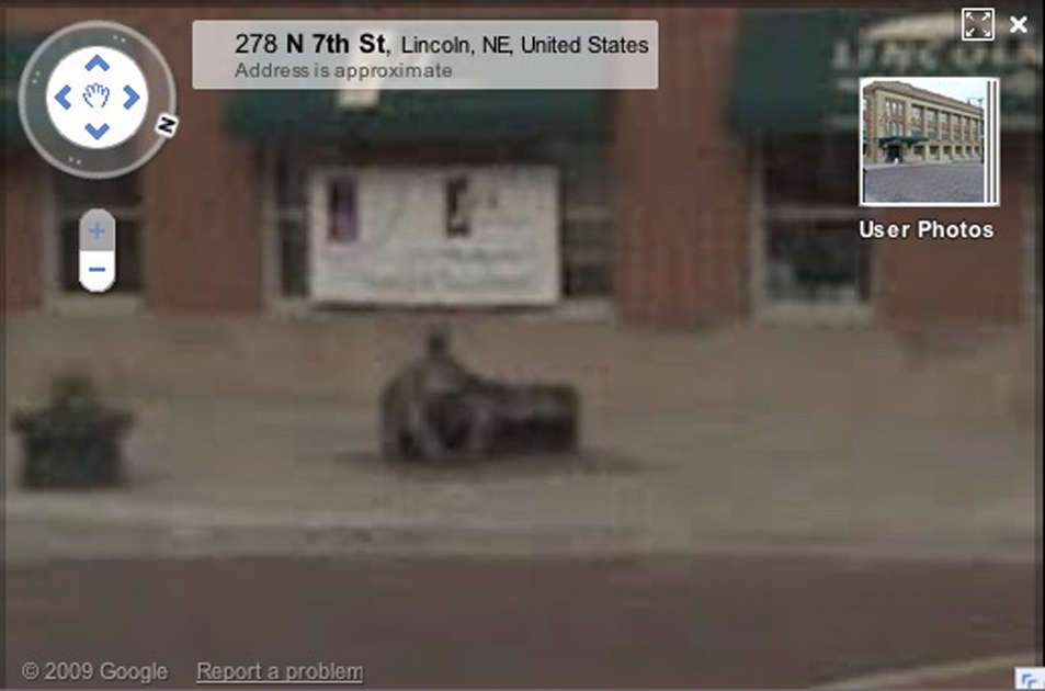 Blurry, zoomed in screenshot of Street View in Lincoln, Nebraska, showing a statue of a man sitting on a bench