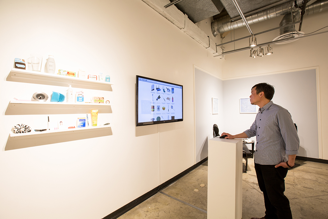 A person in a gallery is using a mouse on a pedestal to navigate this website on a wall-mounted monitor. Next to the monitor are three small shelves with some of the actual objects in the list.